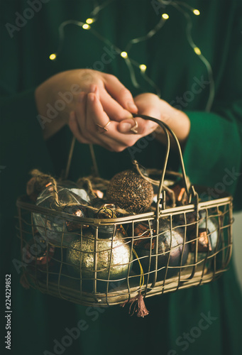 Wire basket full of Christmas or New Year tree vintage decoration toys in hands of woman in green festive dress, holiday garland lights bokeh at background, selective focus, vertical composition photo