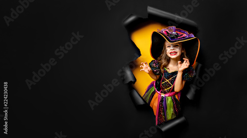 Happy Halloween. Laughing funny child girl in a witch costume of halloween looking, smiling and scares through a hole of black and yellow paper background. Copyspace