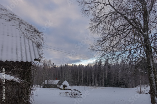 winter morning in the countryside, the sky is cloudy before snowfall, everything is covered with snow, the old barn, the hay in the canopy and the metal wheels are shown below.