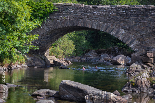 Father and daughter swimming in the River Duddon by old stone bridge in Ulpha in the Lake District National Park, UK. Scenic view of English countryside on a sunny summer day.