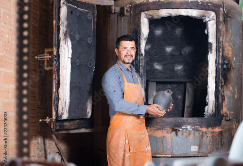 mature man potter holding black glazed ceramic vessel next to kiln photo