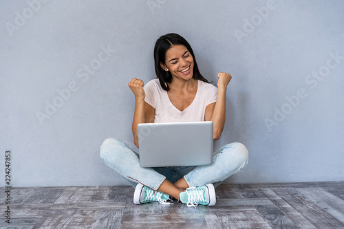 Portrait of excited young casual girl celebrating success while sitting with laptop computer isolated over gray background.