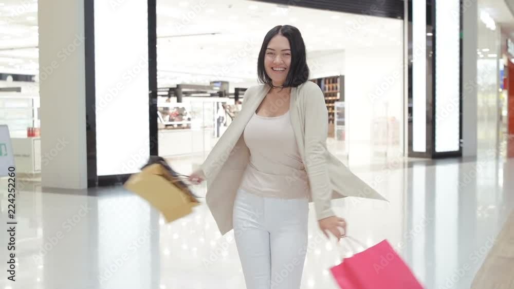 Woman in soft colored jeans and jacket walking and spinning at the mall while carrying shopping bags in both hands. Woman in soft colored jeans and jacket walking and spinning at the mall while carrying shopping bags in both hands.