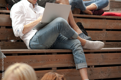 modern fashionable guy sits on a wooden bench among people in the park and works on a laptop