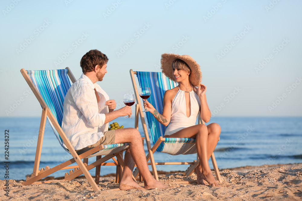 Young couple with glasses of wine in beach chairs at seacoast