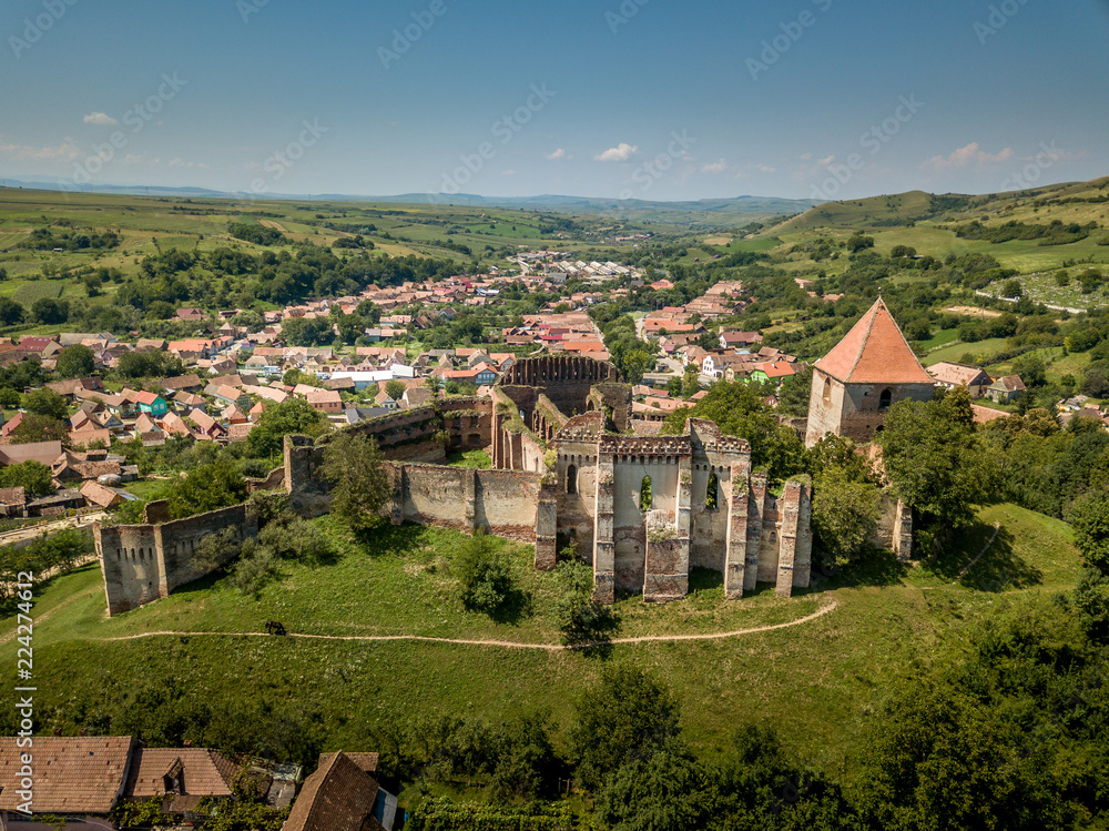 Aerial view of ruined Gothic Saxon medieval Slimnic castle near Sibiu, Romania with donjon, church, barbican, walls on a green hill with blue cloudy sky