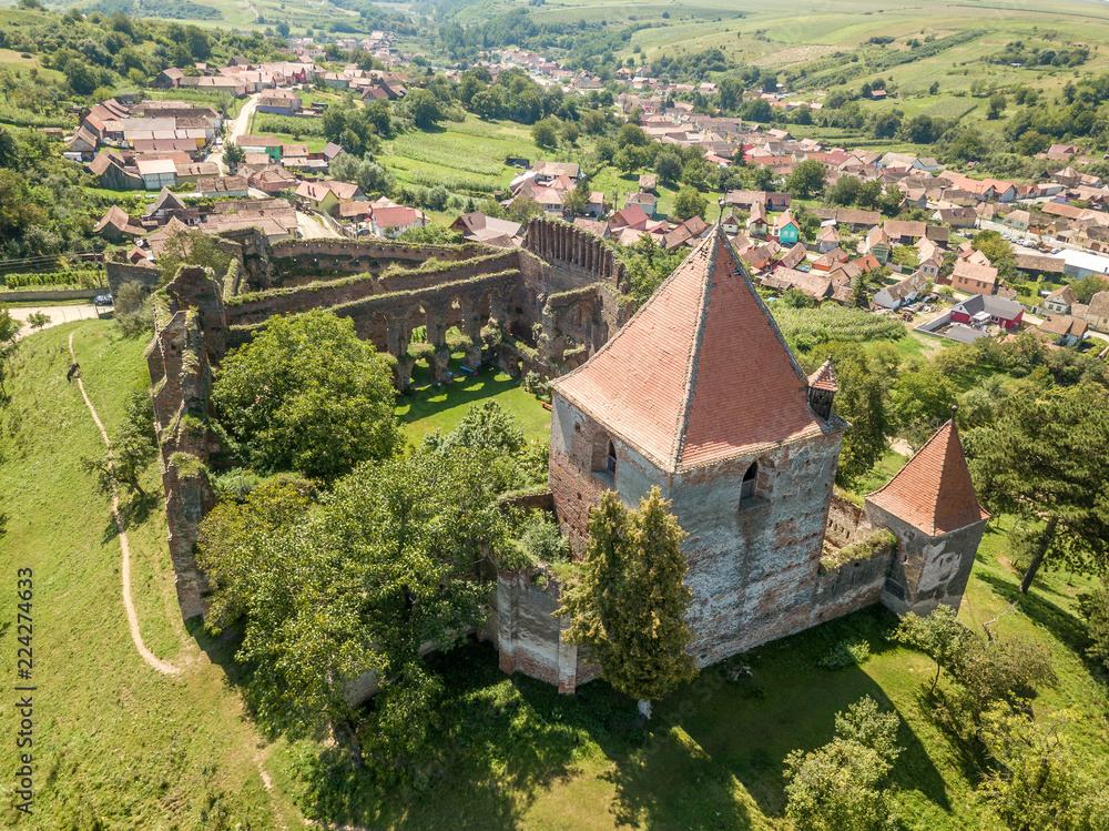 Aerial view of ruined Gothic Saxon medieval Slimnic castle near Sibiu, Romania with donjon, church, barbican, walls on a green hill with blue cloudy sky