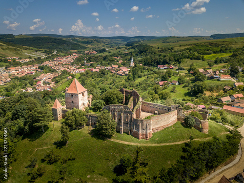 Aerial view of ruined Gothic Saxon medieval Slimnic castle near Sibiu  Romania with donjon  church  barbican  walls on a green hill with blue cloudy sky