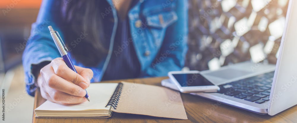 A woman's hand is writing on a notepad with a pen and a phone with a laptop.Web banner.