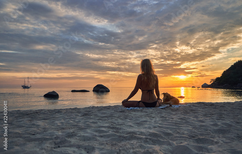 Young caucasian woman with dog meditating on the beach and watching dawn