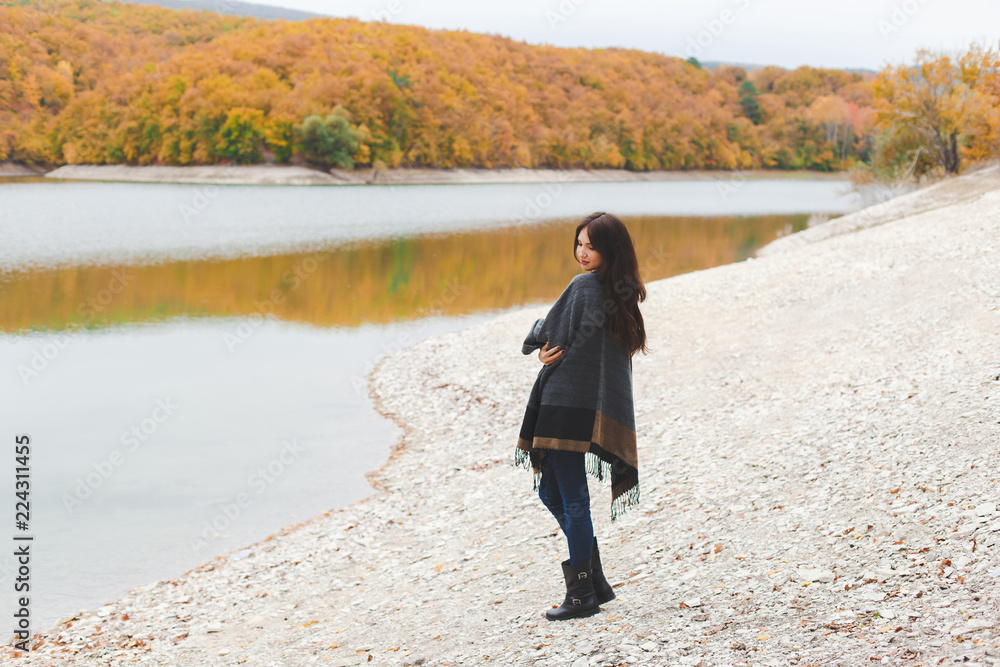 Young brunette woman wearing grey poncho standing alone near the lake in autumn. Weekend outdoors, cloudy cold weather, fall colors.