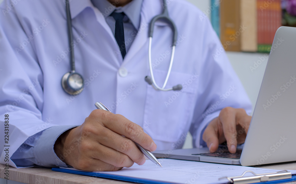 Doctor with stethoscope working on laptop computer and writing note on paperwork in doctor room at clinic or hospital.