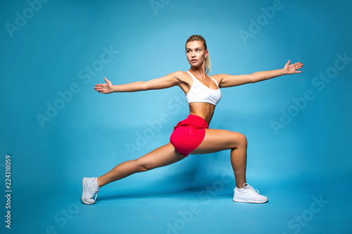 young sports happy sexy blonde girl in red skirt and white top is doing yoga exercise and looking straight with hands to the sides on the blue wall background