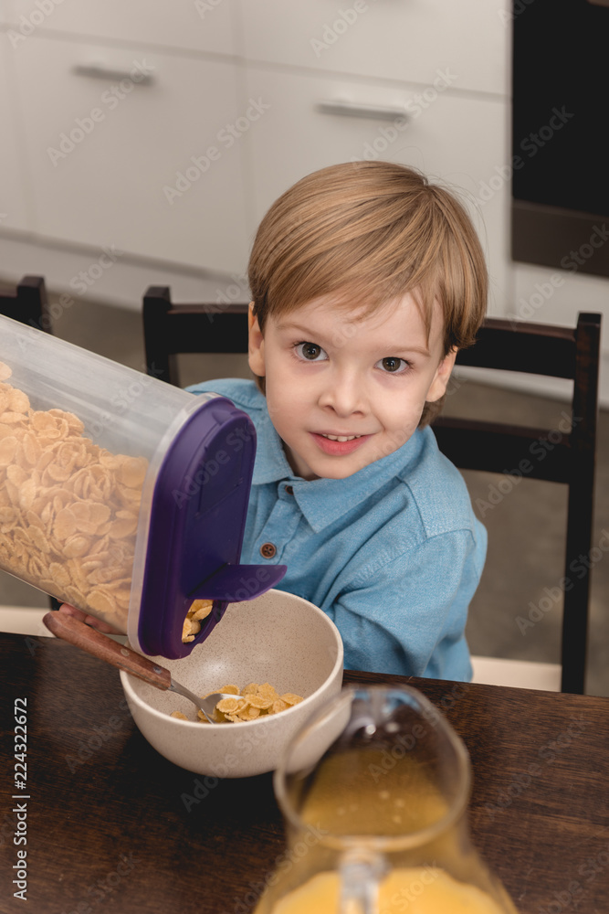 adorable little kid pouring cereal in bowl for breakfast and looking at camera adorable little kid pouring cereal in bowl for breakfast and looking at camera