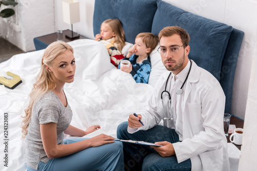 high angle view of pediatrician and mother looking at camera while sick kids lying in bed on background