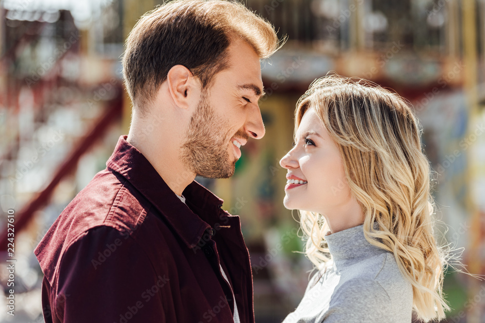 portrait of affectionate couple in autumn outfit looking at each other near carousel in amusement park