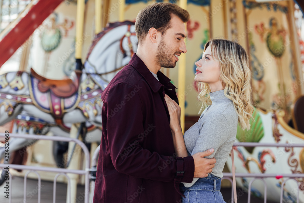 side view of romantic couple in autumn outfit cuddling near carousel in amusement park side view of romantic couple in autumn outfit cuddling near carousel in amusement park