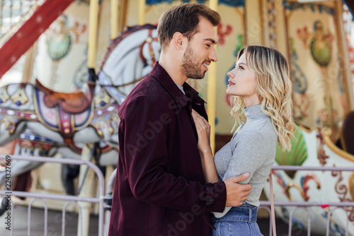 side view of romantic couple in autumn outfit cuddling near carousel in amusement park