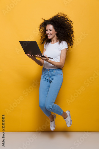 Full length photo of european woman 20s wearing casual clothes smiling and jumping while holding black laptop, isolated over yellow background