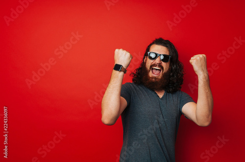 Cheerful bearded man with long curly hair wearing sunglasses and celebrating over red background