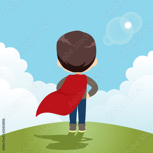 Back view of cute little super hero boy standing on high mountain with big blue sky and waving red cape