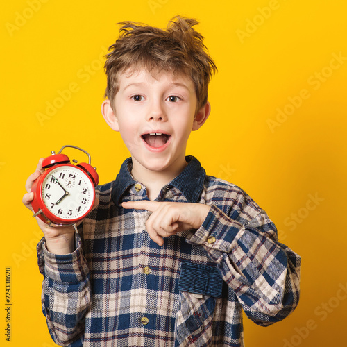 Cute little boy with alarm clock,isolated on yellow. Funny kid pointing at alarm clock at 7 o'clock at morning. Excited boy overslept before school. People, school, time and lifestyle concept.
