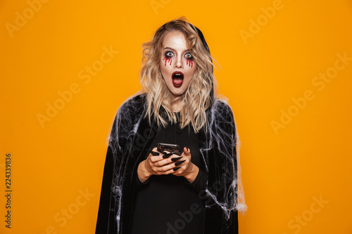 Blond woman wearing black costume and halloween makeup holding mobile phone, isolated over yellow background