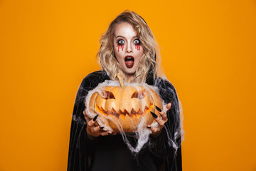 Magician woman wearing black costume and halloween makeup holding carved pumpkin, isolated over yellow background
