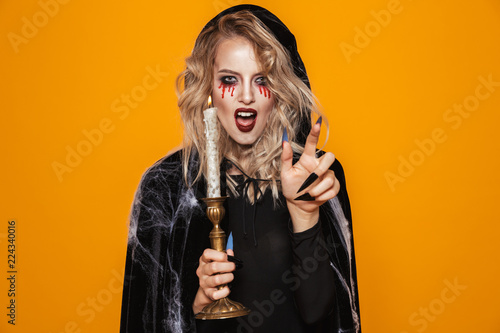 Horror wizard woman 20s wearing black costume and halloween makeup holding candle, isolated over yellow background