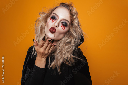 Frightening woman wearing black costume and halloween makeup looking at the camera, isolated over yellow background