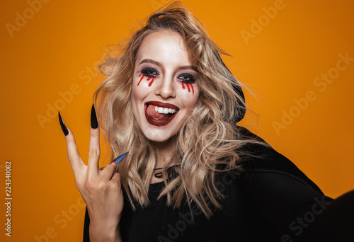 Joyful witch woman wearing black costume and halloween makeup smiling at the camera, isolated over yellow background