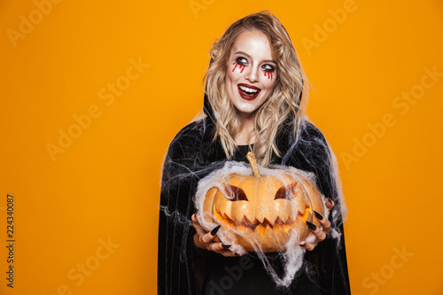 Scary woman wearing black costume and halloween makeup holding carved pumpkin, isolated over yellow background