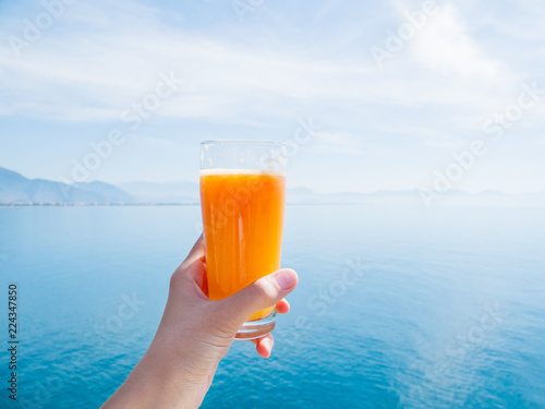 Hand with fresh tasty freshly-squeezed juice of ripe oranges in glass. Calm sea with mountains on the horizon. Kemer, Turkey.