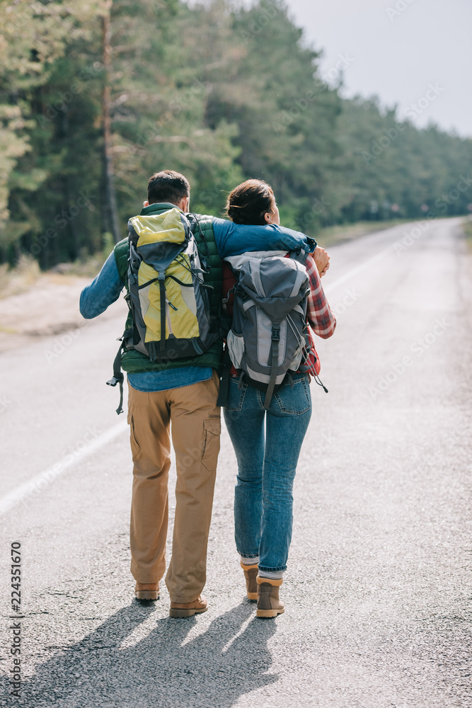 back view of couple of travelers with backpacks walking on road