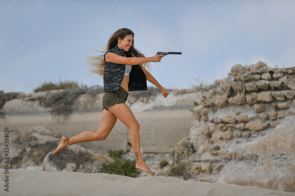 Beautiful girl running and jumping with a gun on a sandy sea hill with