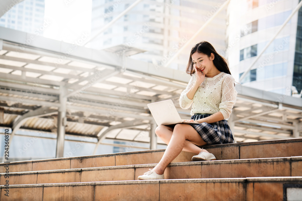 Portrait of a beautiful Asian teenage girl Sitting play on laptop stairs in the big city Looking for learning information. Researching a variety of information. Including online shopping for women.
