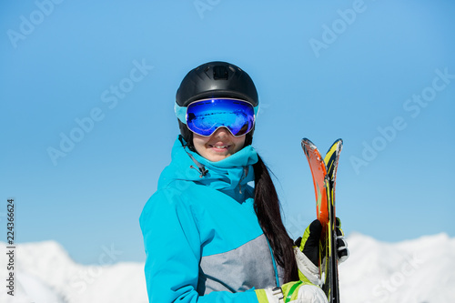 Photo of young smiling female athlete in helmet with skis in hand against blue sky and snowy hill