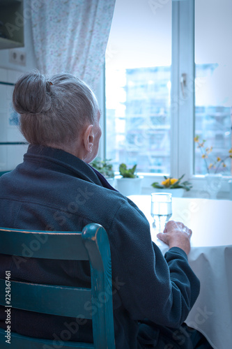 Loneliness concept, old-age depression: old woman sitting alone at her kitchen table looking out of the window, blue filter effect.