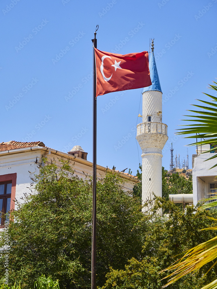 Minaret of Kelerlik Mahallesi Cami mosque with the official flag of the Republic of Turkey waving in the foreground. Bodrum, Mugla Province, Turkey.