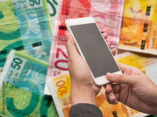 Slika na platnu Woman holding white smart phone with blank desktop screen on blur israeli shekel money background