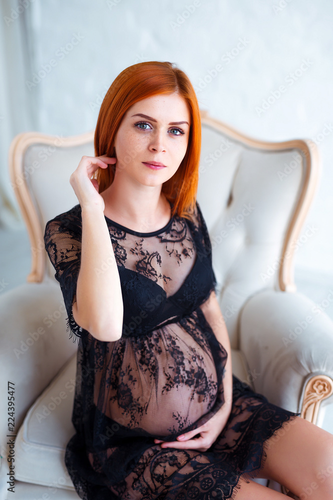 Fototapeta premium Pregnancy beauty. Beautiful elegant readhead pregnant woman in charming black dress posing in tender home interior.