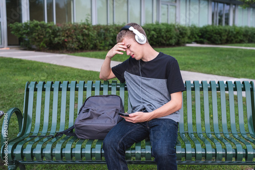 Depressed teenager sitting on bench outside of his school while listening to music with his headphones. © Brian