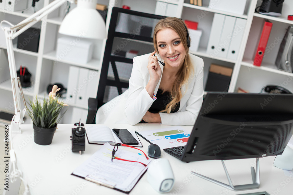 A young girl in headphones with a microphone sits at a table in the office and looks at the monitor. A young girl in headphones with a microphone sits at a table in the office and looks at the monitor.