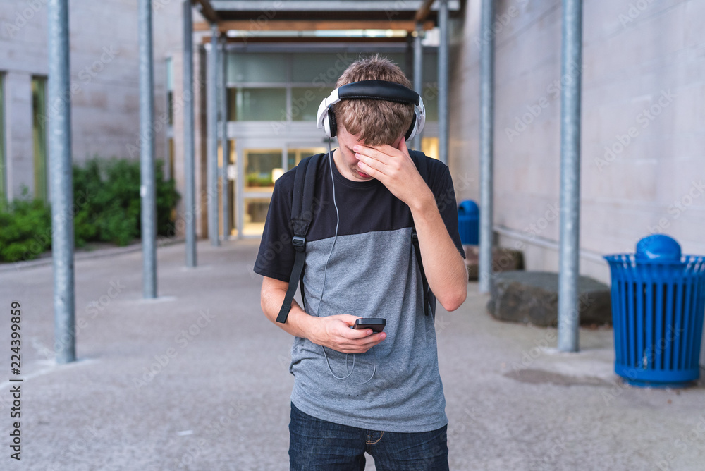 Sad teen covering his face while standing in front of his school and listening to music.