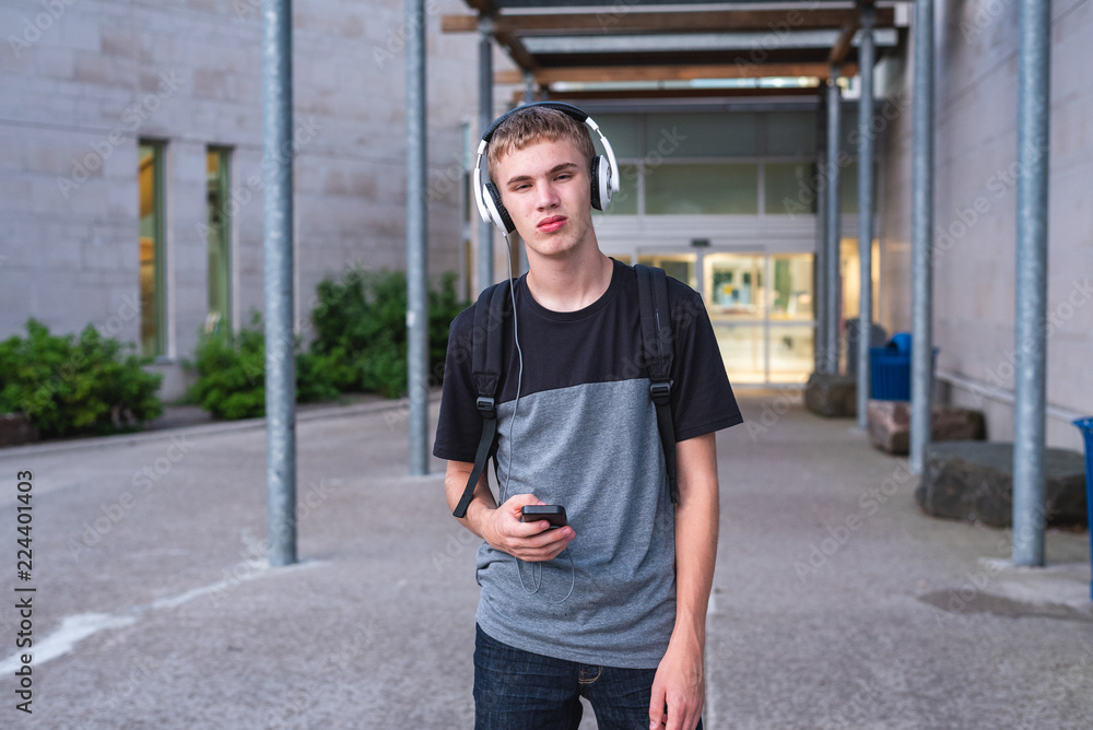 Sad teenager standing in front of his school while listening to music with his headphones.