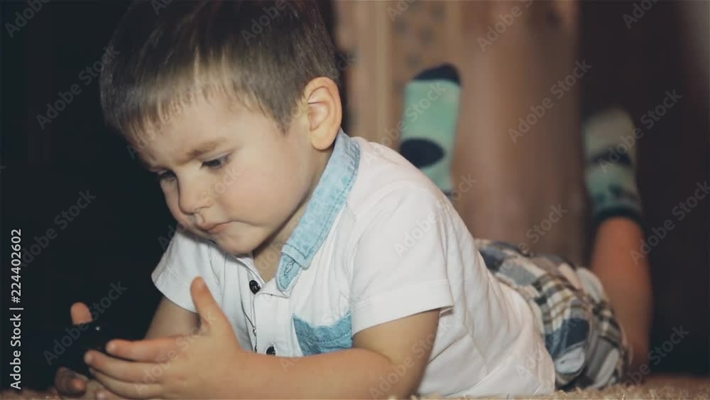 The boy lies on the floor and carefully examines the mobile phone The boy lies on the floor and carefully examines the mobile phone
