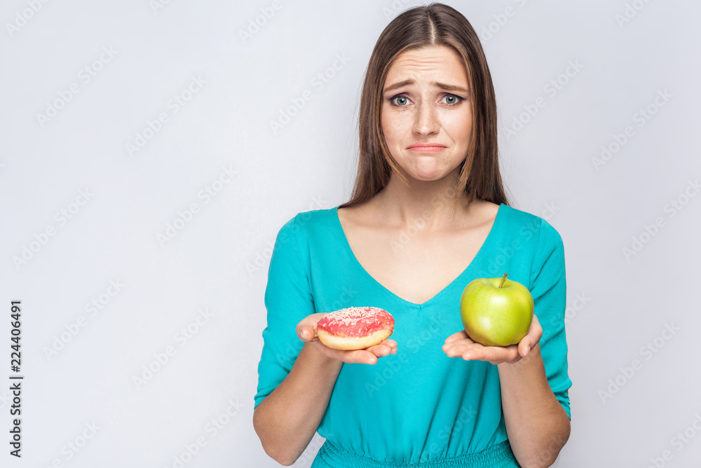 Portrait of upset dreaming young beautiful girl in blue blouse standing, holding pink donut and green apple in the hands like weighing with unhappy face on grey background. Indoor, isolated,copy space Portrait of upset dreaming young beautiful girl in blue blouse standing, holding pink donut and green apple in the hands like weighing with unhappy face on grey background. Indoor, isolated,copy space