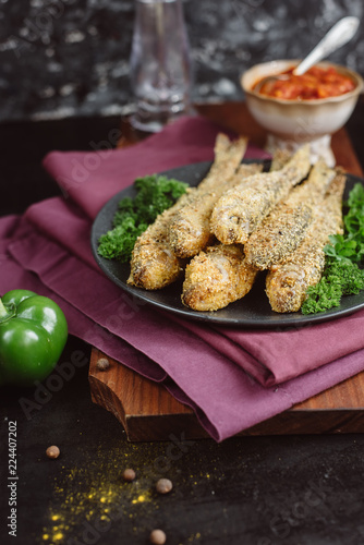 Fried fish in a plate with tomato sauce, greens and peppers on wooden board and a dark background