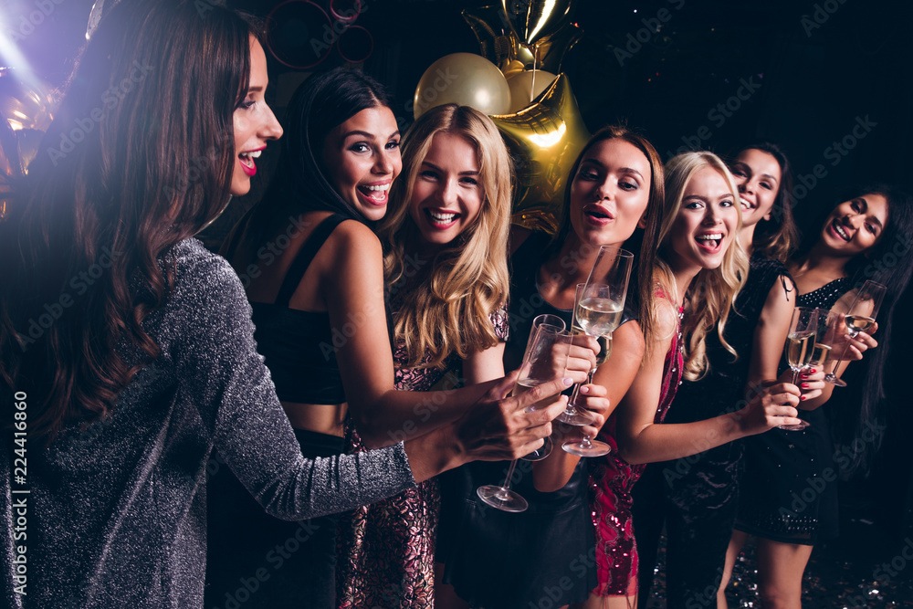 Pure celebration. Beautiful young women in evening gown holding champagne glasses and looking at camera with smile while celebrating in nightclub - obrazy, fototapety, plakaty 