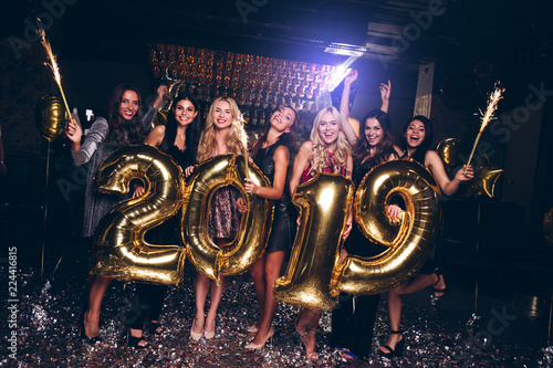 Happy new year! Beautiful young women in evening gown holding balloons and looking at camera with smile while celebrating in nightclub